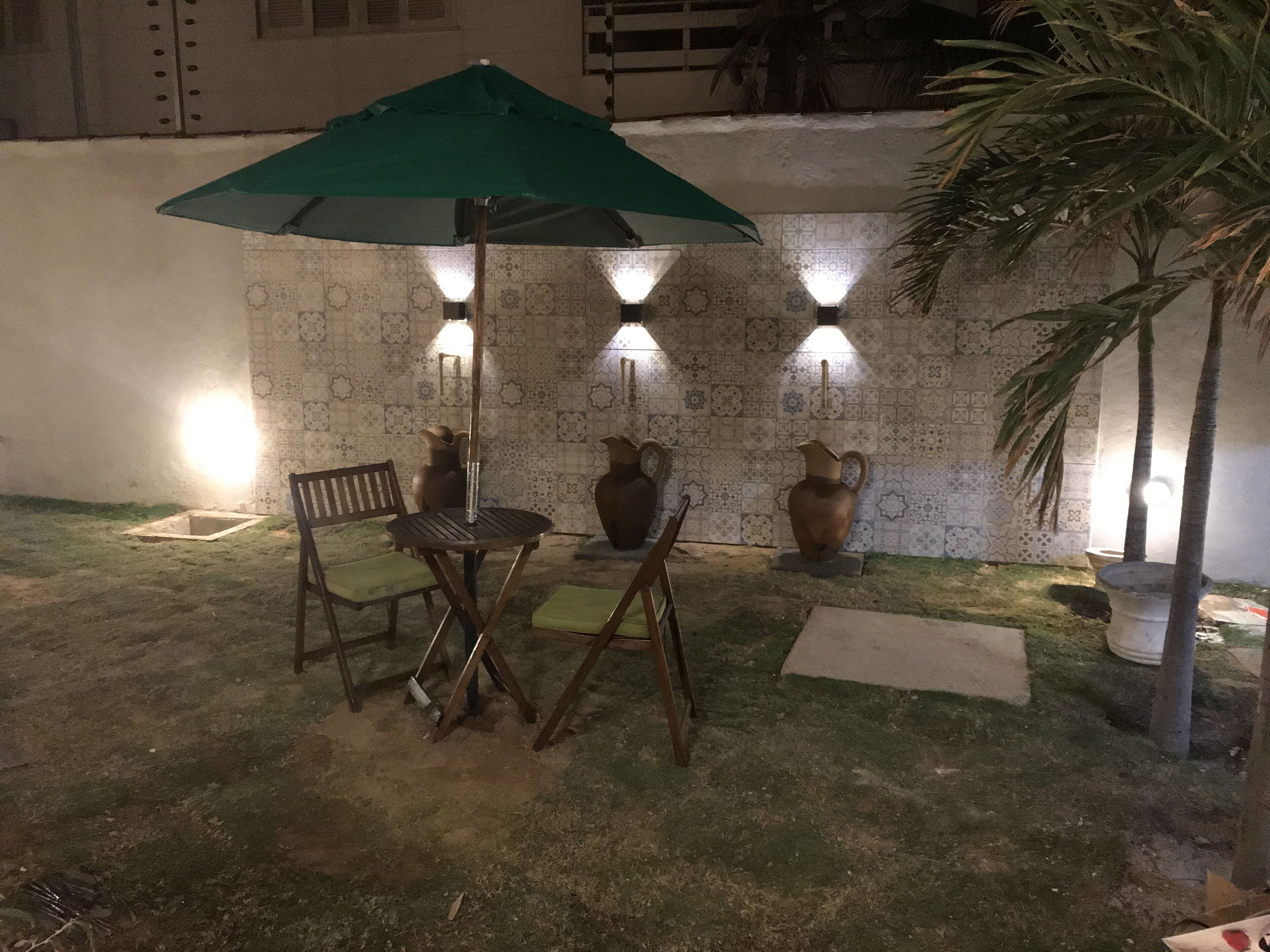 An outdoor seating area with a table and chairs under a green umbrella, decorative vases, and wall-mounted lights.