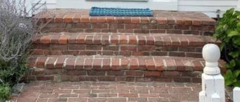 Een wit gebouw met een blauwe deur, bakstenen trappen en een witte hekken.'