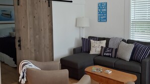 A modern living room with a dark grey sofa, two armchairs, a wooden coffee table, and a sliding barn door. A blue sign with text is on the wall, and a window with blinds is visible. - Spacious 1 Bedroom, First Fl., Beach Access, in quiet East End. 
 (Provincetown)