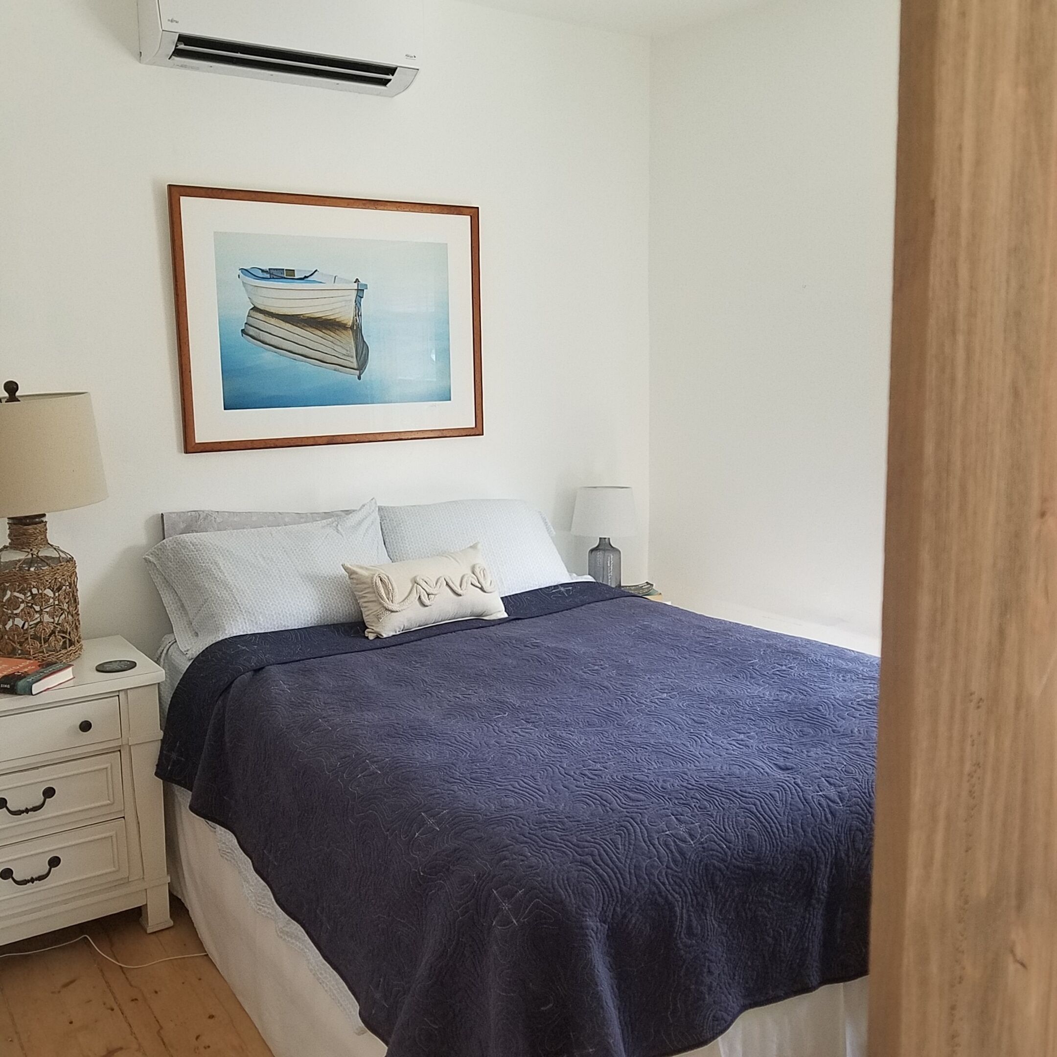 Une chambre à coucher avec un lit, une table de chevet, une lampe, un tableau encadré et un climatiseur.