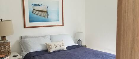 Une chambre à coucher avec un lit, une table de chevet, une lampe, un tableau encadré et un climatiseur.