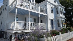 A two-story white house with a balcony, a flag, and a garden. - Spacious 1 Bedroom, First Fl., Beach Access, in quiet East End. 
 (Provincetown)