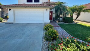 A single-story house with a white garage door, a red door, and a small front yard with a palm tree and flower beds. - Charming Southwest Style Vacation Home  (Phoenix)