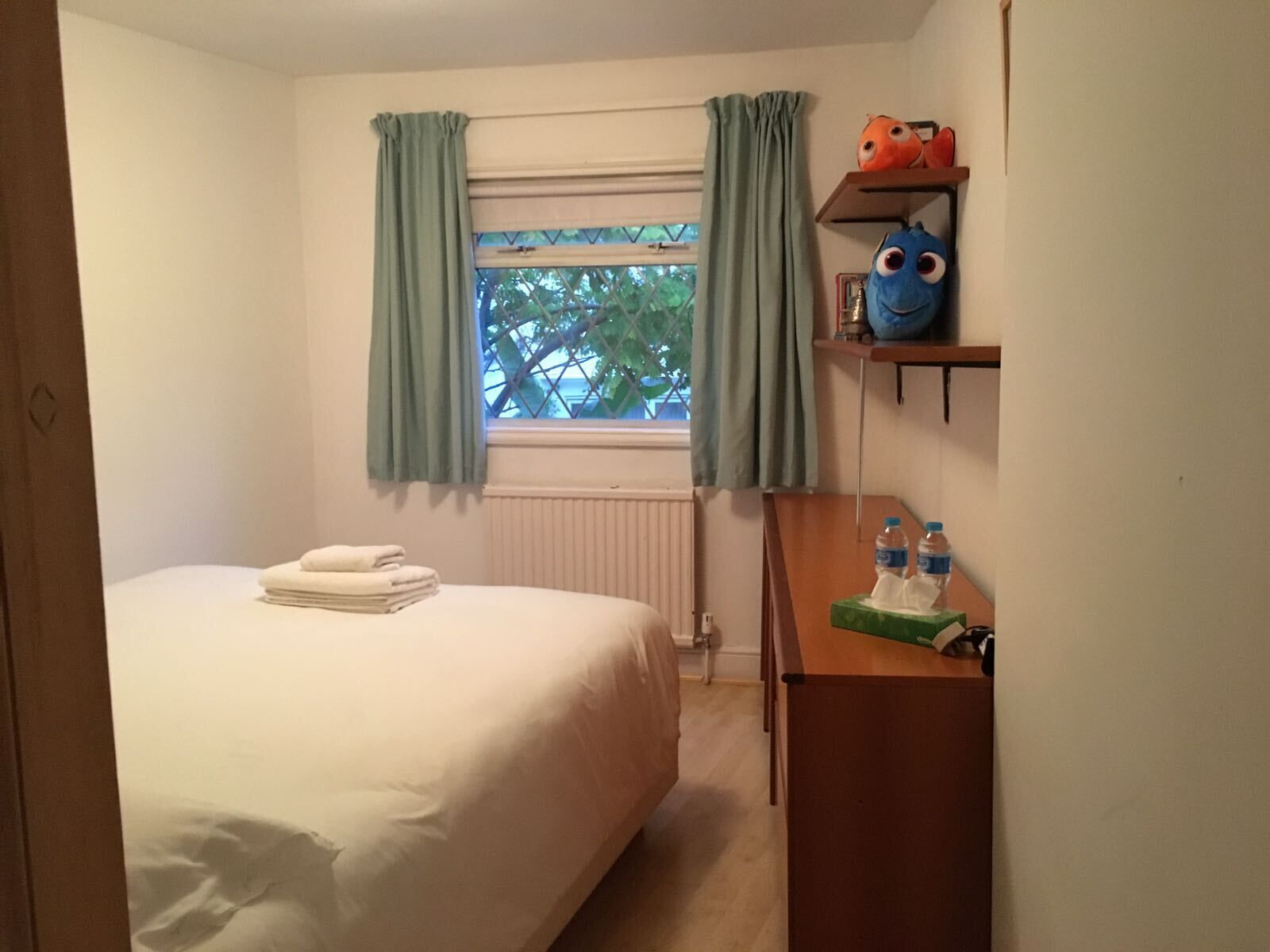 Un dormitorio con cama, un radiador, una ventana con cortinas, una estantería con peluches y una mesita de noche con pañuelos de papel y botellas de agua.