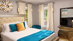 A hotel room with a tufted headboard, a bed with white and blue bedding, teal pillows, and a blue throw blanket. There is a television on a gold-framed stand, and a decorative wall piece above the bed. - Atlanta Pool Home Near Airport, Near Airport 8 guests Max (Atlanta)
