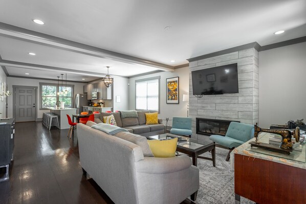 A modern living room with a grey sofa, a blue armchair, and a fireplace. There is a kitchen in the background with grey cabinets and a dining area.