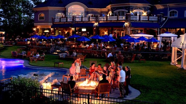 A large outdoor dining area with multiple tables and blue umbrellas, a pool with a fire feature, and a building with multiple balconies. - 10 mins. to downtown Saratoga and SPAC. 1,000 sq. ft. Spacious and quiet. (Ballston Spa)