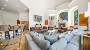 Amplia sala de estar con un sofá seccional gris grande, una mesa de centro y una chimenea. Hay sillones de mimbre y una zona de comedor con una mesa de vidrio.