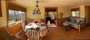 A dining area with a round table, chairs, and a chandelier. There is a sofa and a lamp in the adjacent living area.