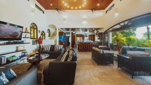 Un salón amplio con un televisor grande, asientos cómodos, una cocina al fondo y un balcón con vistas.