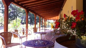 Un patio al aire libre con sillas de metal y una mesa redonda, rodeado por una pérgola de madera y una pared amarilla.