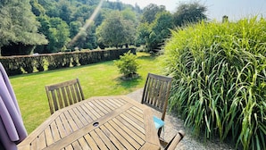 A wooden table with chairs, a small blue cushion, and a view of a lush garden with a hedge and tall grass. - Le Gite De Parc (2 Bedrooms, Sleeps 4) (Souleuvre En Bocage)