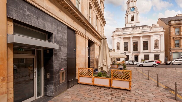 L’entrée d’un hôtel, avec une porte moderne en verre, un trottoir pavé de briques et un bâtiment historique surmonté d’une tour horlogère en arrière-plan.