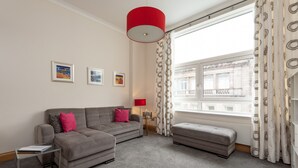 Un salon avec un canapé gris, une petite table d’appoint et une lampe rouge. Trois tableaux encadrés ornent le mur, et une fenêtre est agrémentée de rideaux à motifs.