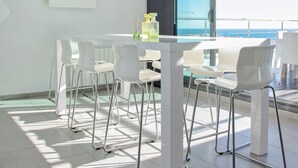 Un espace de repas moderne avec une table et des chaises blanches, un luminaire suspendu et une vue sur l’océan grâce à de grandes fenêtres.