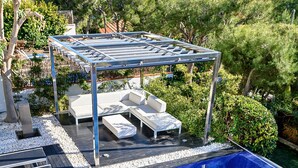 Une piscine avec une terrasse en bois, des chaises longues et une pergola agrémentée de coussins.