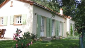 Une maison à deux étages avec des volets verts, un jardin fleuri et un banc en bois.