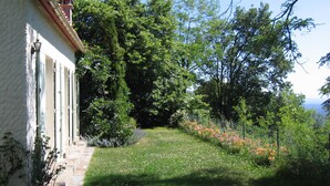 Une maison avec une façade en pierre, un toit en tuiles et un jardin où poussent différentes plantes et fleurs.