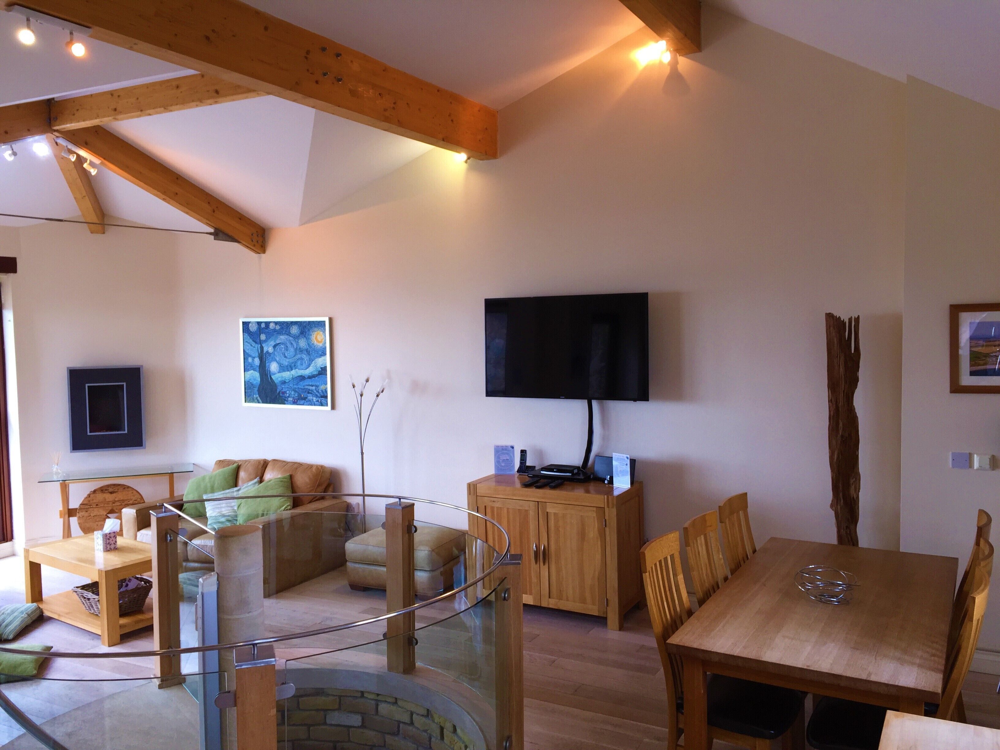 A spacious living area with a glass staircase, wooden furniture, a flat-screen TV, and artwork on the walls.