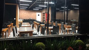 Uma ampla área de refeições em ambiente interno, com diversas mesas e cadeiras, tetos altos com vigas aparentes e paredes de tijolo.