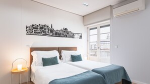 A hotel room with a bed, bedside tables, a cityscape wall art, a window with a view, and an air conditioning unit. - Feel Porto Downtown City Roofs (Oporto)