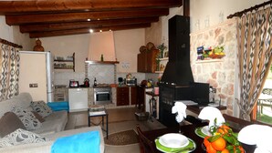 Een woonkamer met een bank, een eettafel gedekt met borden en een fruitschaal, een keuken met apparatuur, en een open haard.
