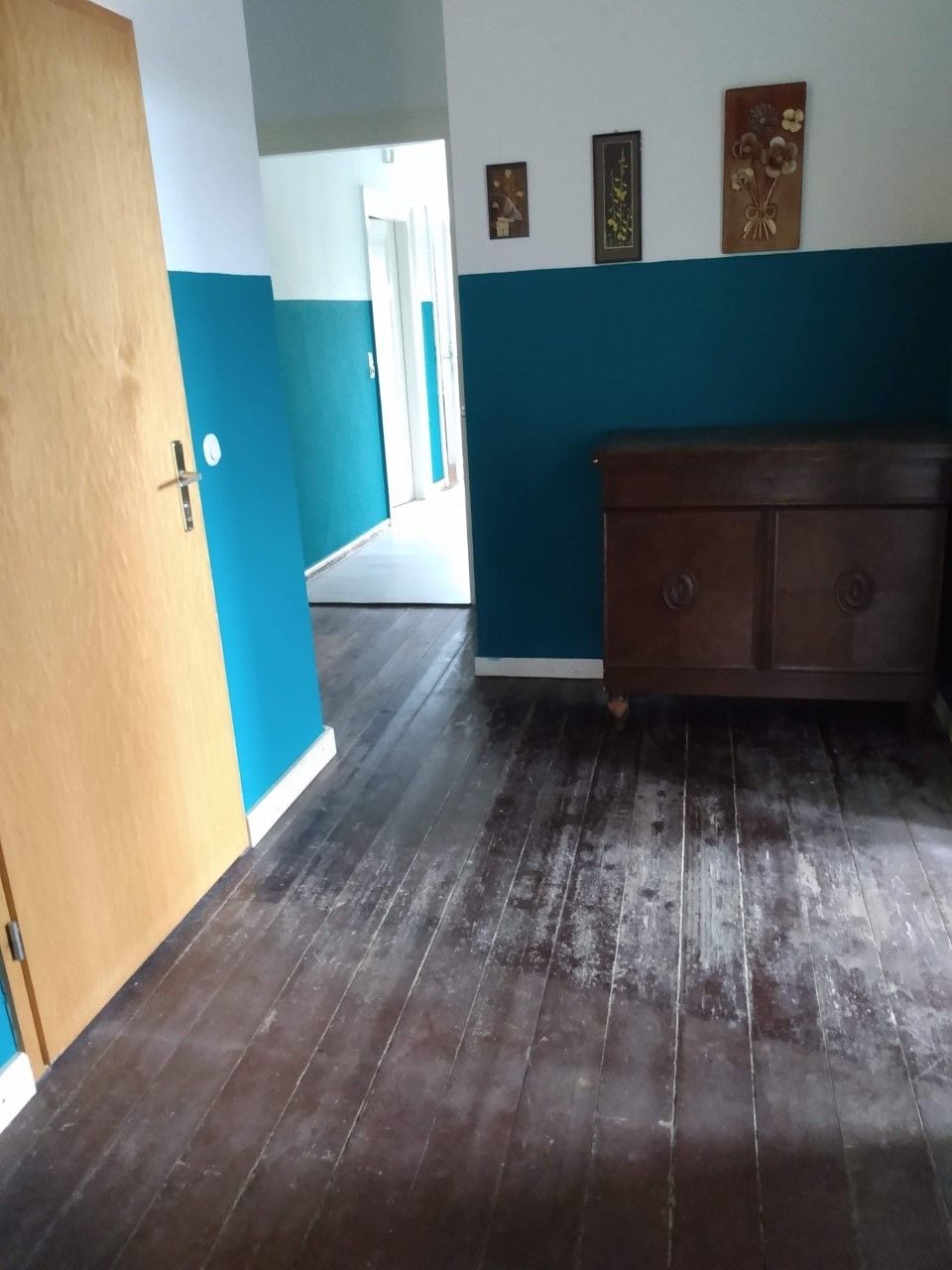 A hallway with dark wooden flooring, a wooden door on the left, and a blue wall with three framed pictures.