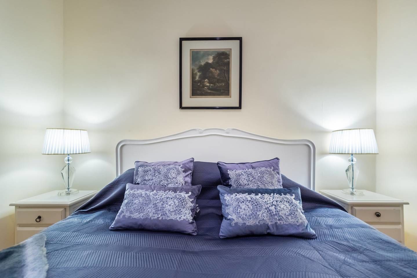 A neatly made bed with a blue bedspread and four pillows, two nightstands with lamps, and a framed picture on the wall.