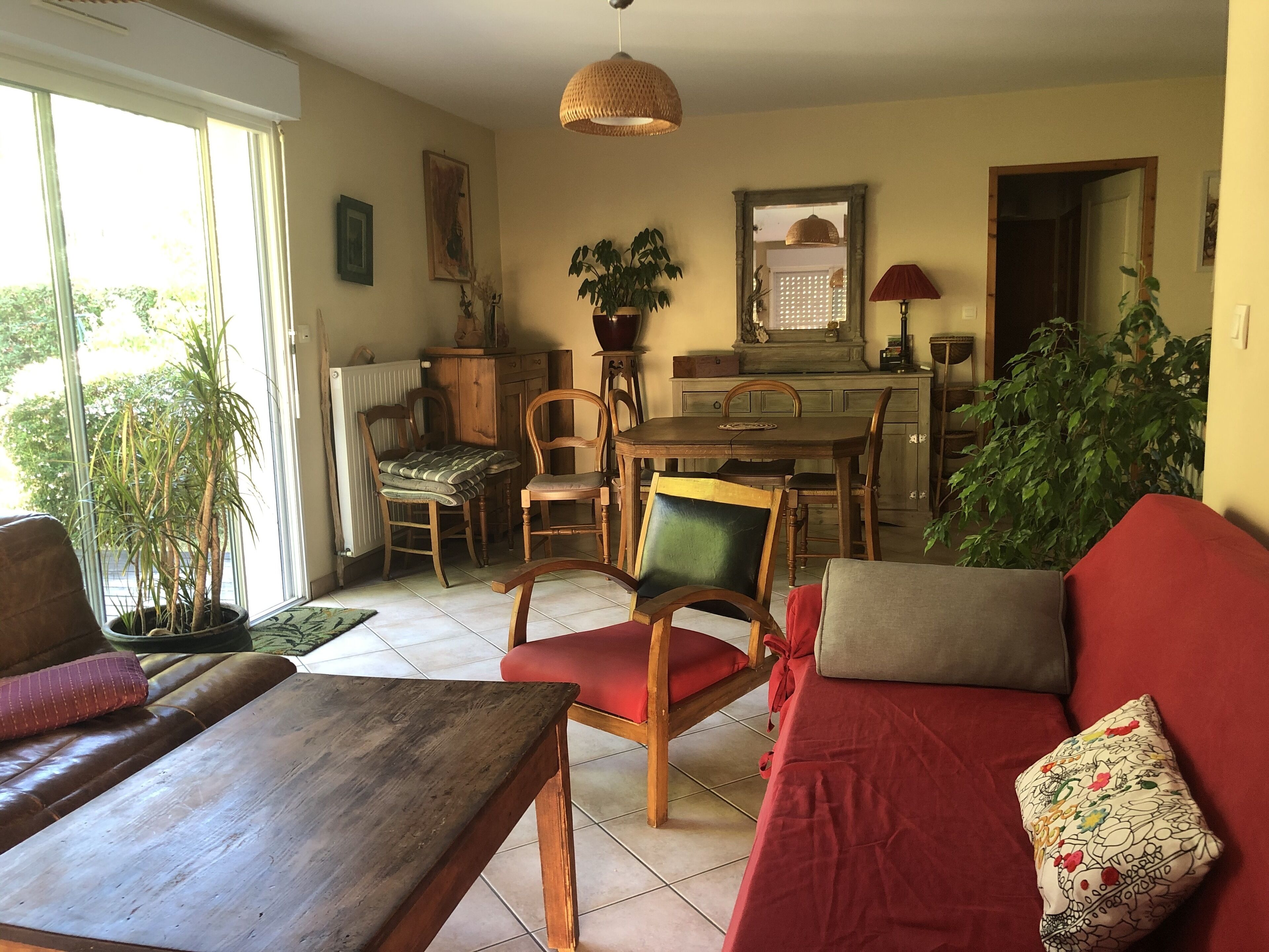A cozy living room with a wooden coffee table, a red sofa, and a dining area with a wooden table and chairs. There is a large window providing natural light, and a potted plant is visible in the corner.