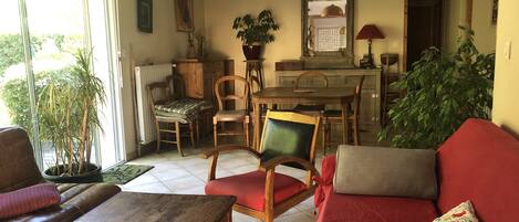 A cozy living room with a wooden coffee table, a red sofa, and a dining area with a wooden table and chairs. There is a large window providing natural light, and a potted plant is visible in the corner.