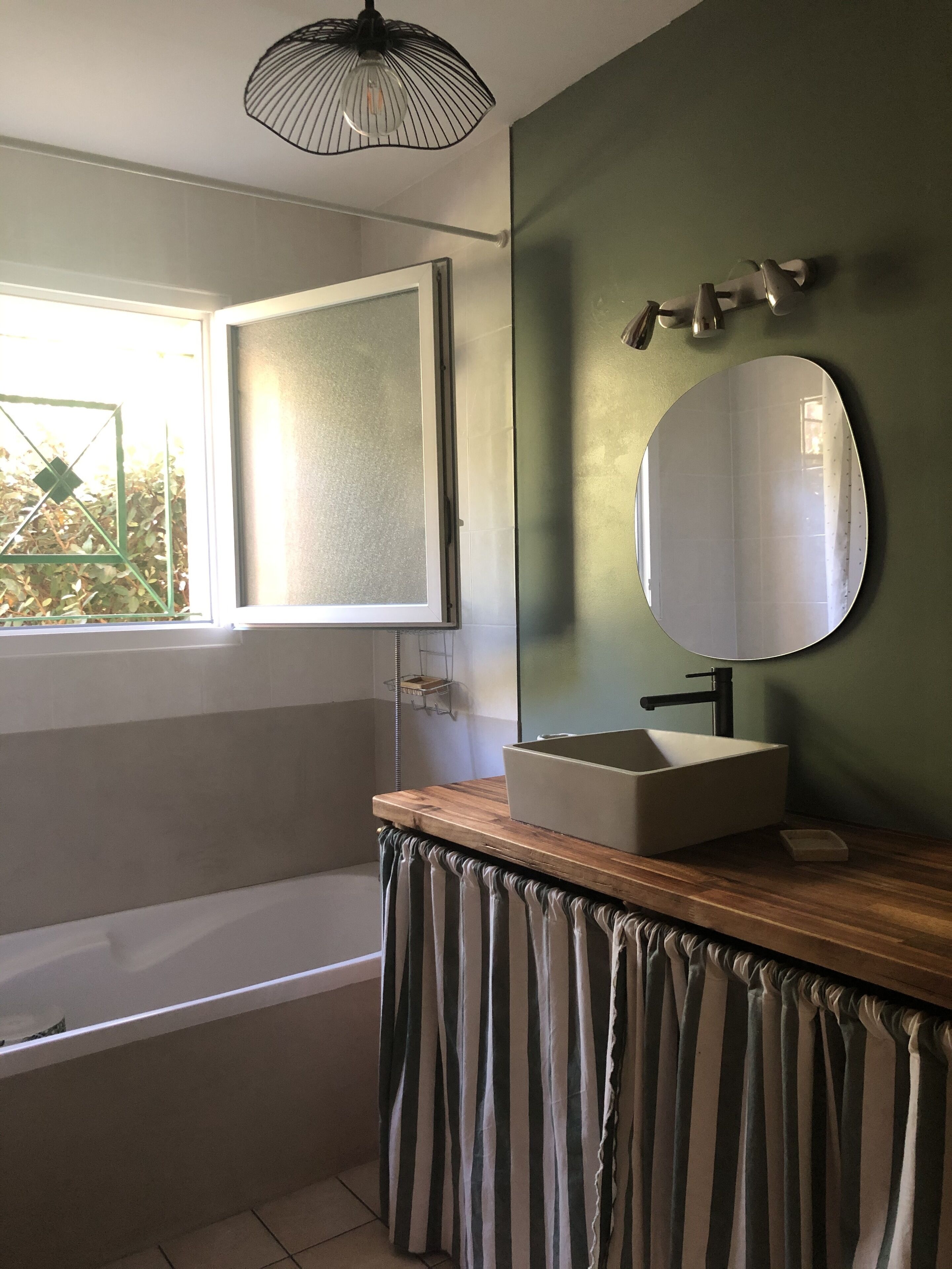 A bathroom with a bathtub, a wooden vanity with a sink, a mirror, and a window with a view of greenery.
