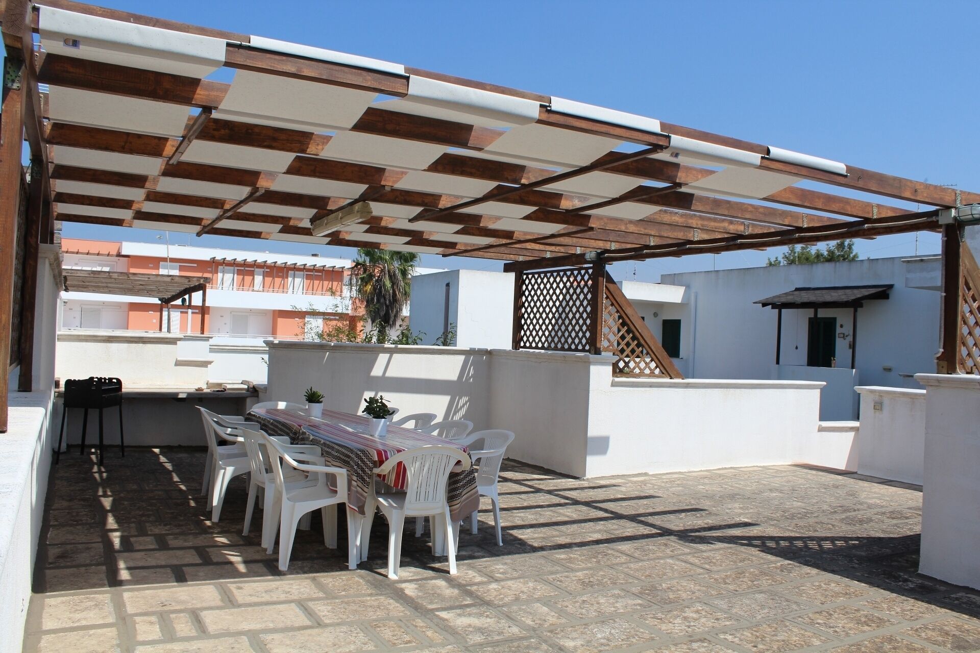 Un patio al aire libre con una mesa y sillas, una pérgola arriba y vistas de edificios al fondo.