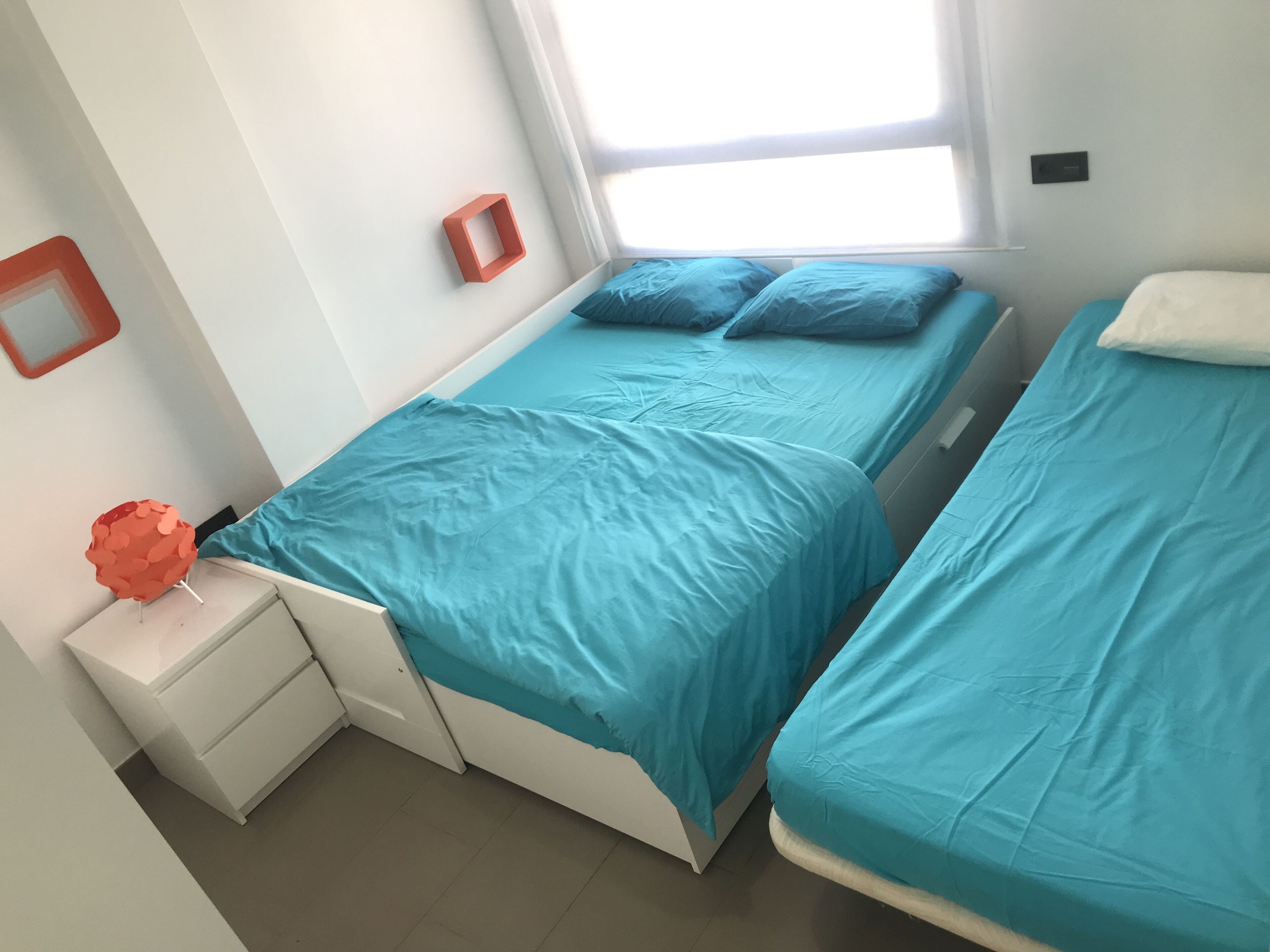 Ein Zimmer mit zwei Betten, jeweils mit blauer Bettdecke und Kissen, einem weißen Nachttisch mit einem roten Gegenstand darauf und einem Fenster mit Vorhängen.