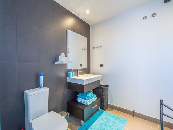 A modern bathroom with a toilet, sink, and mirror. There is a trash bin and a blue towel on a shelf.