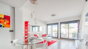 A modern living room with a glass dining table, white chairs, a red bookshelf, and a large window with a view of the outdoors. - Beautiful contemporary Villa (Orihuela)