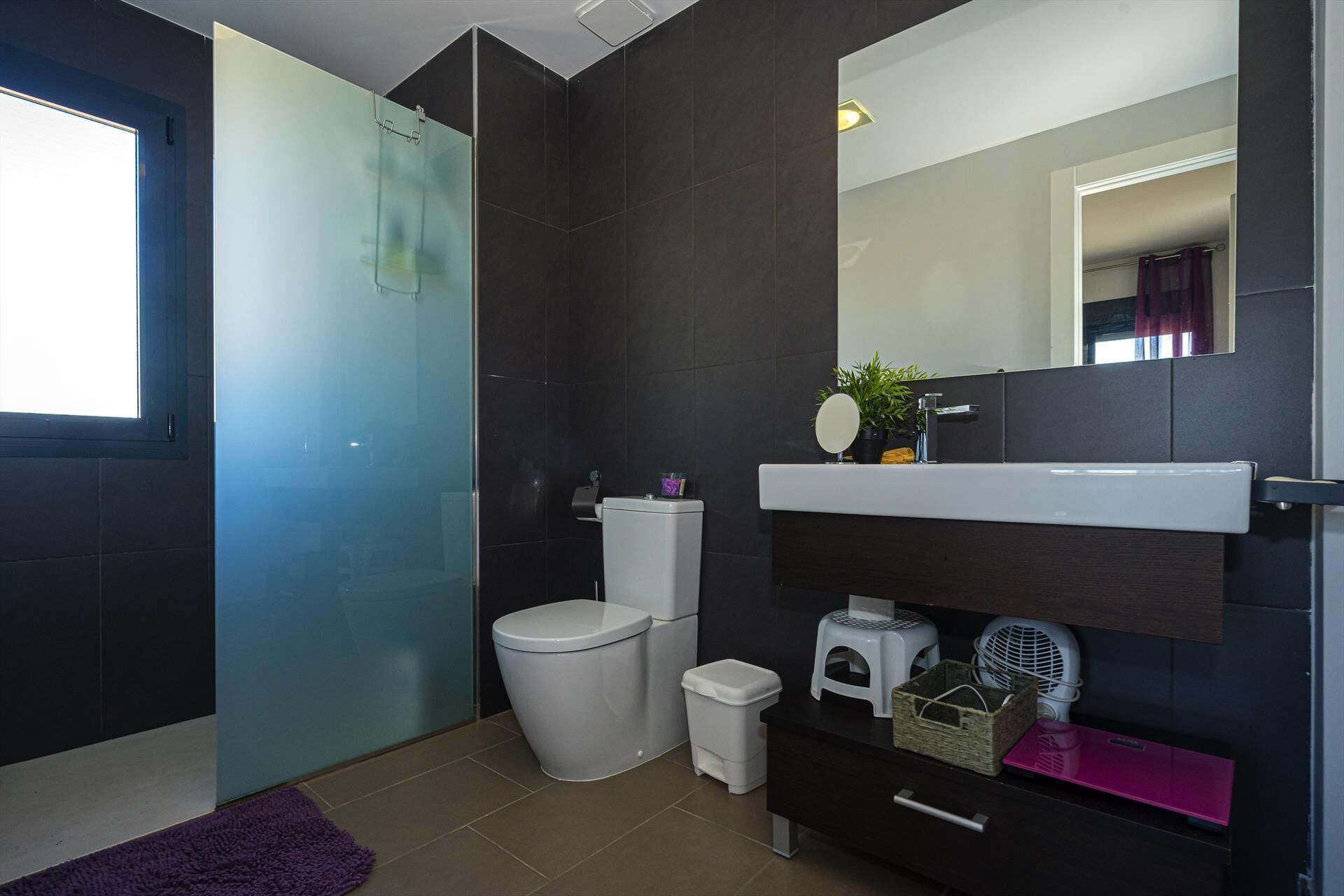 Une salle de bain moderne avec une douche à parois de verre, des toilettes blanches, une petite poubelle, un meuble-lavabo foncé avec un miroir et un panier de rangement rose.