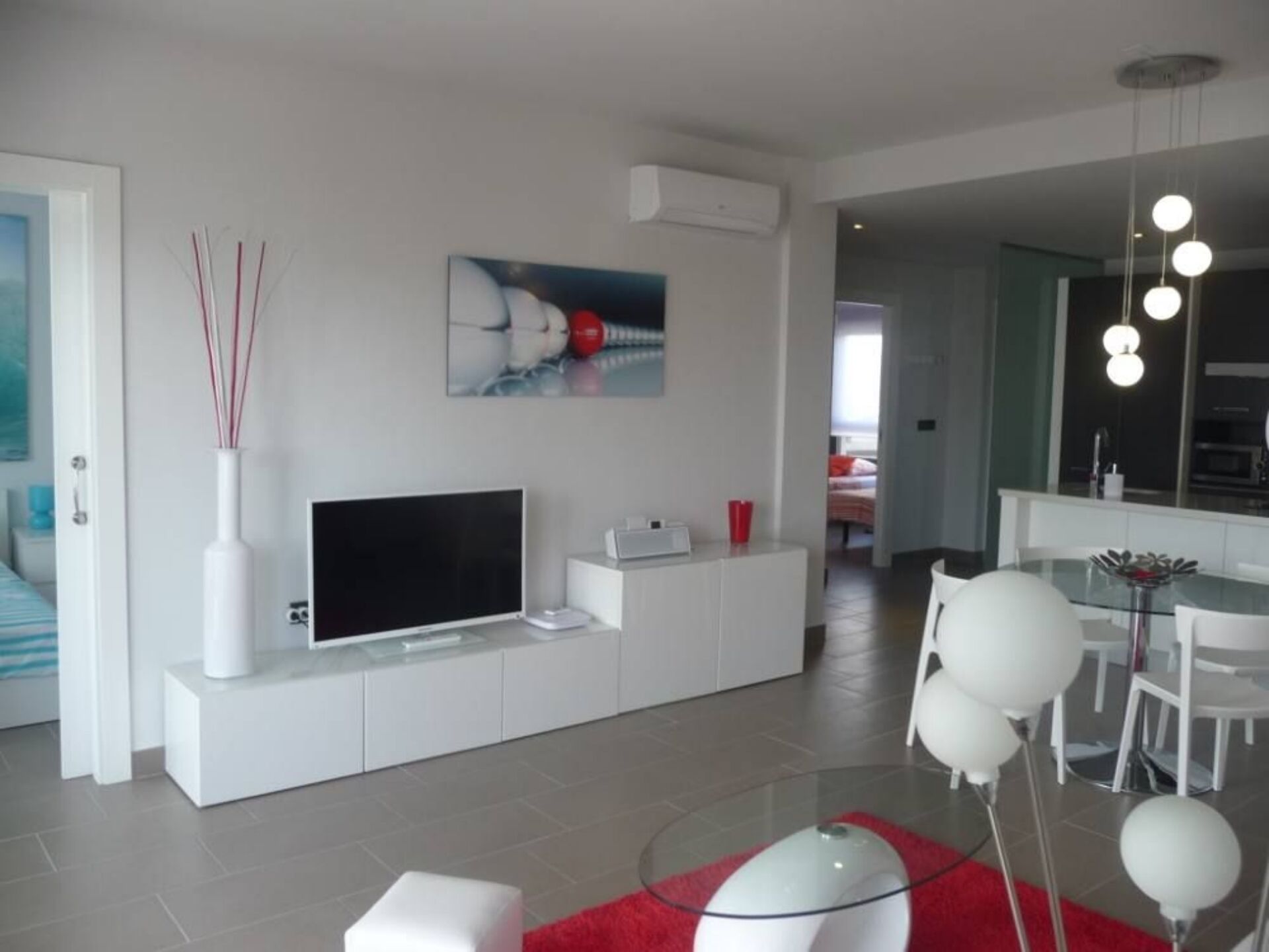 A modern living room with a flat-screen TV, a white entertainment unit, a dining area with a glass table and white chairs, and a red rug.