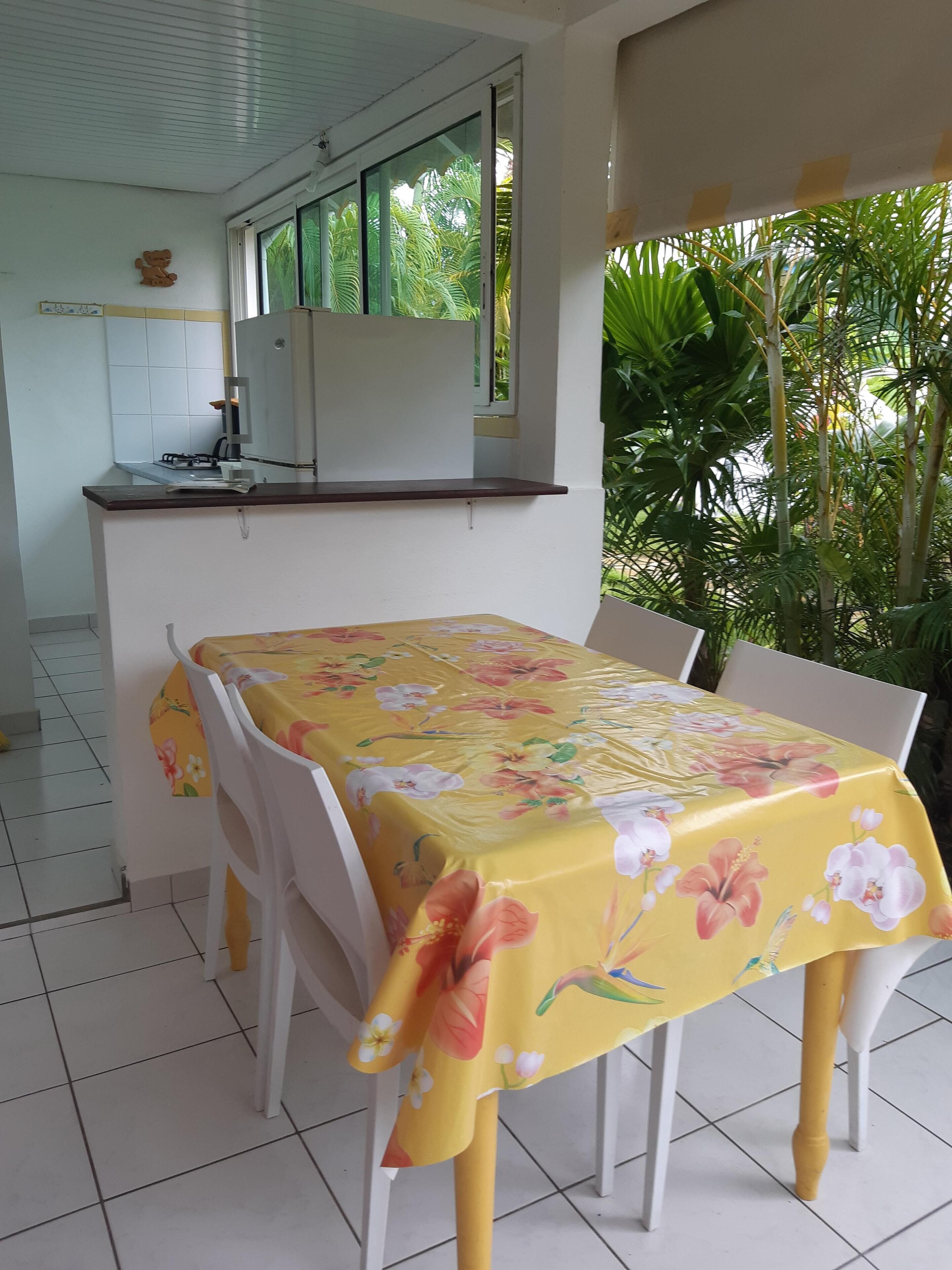 A dining area with a table covered in a floral tablecloth, white chairs, a kitchen area with a gas stove, and a window with a view of greenery.