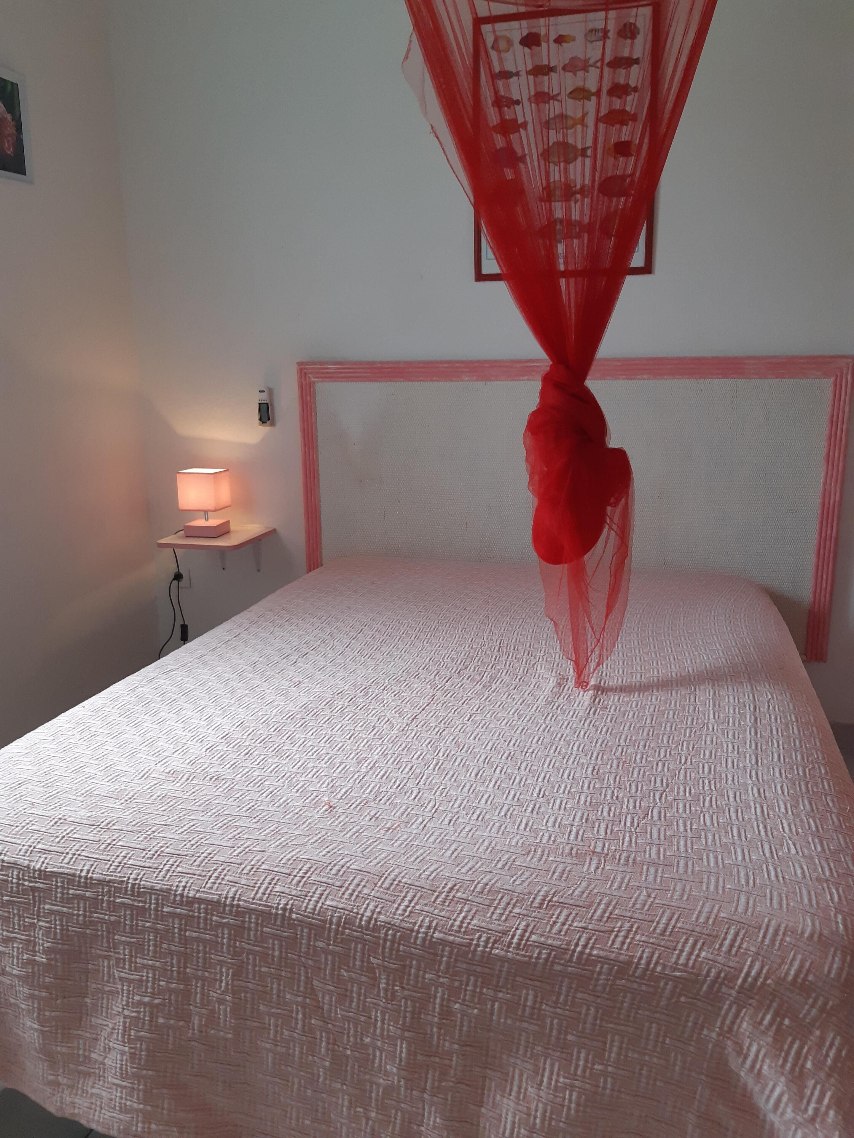 Una cama bien hecha con una colcha estampada, una lámpara de noche y un dosel rojo.