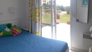 Een slaapkamer met een bed, kleurrijke kussens, een raam met gordijnen, uitzicht op buiten en een wandmodel airconditioner.