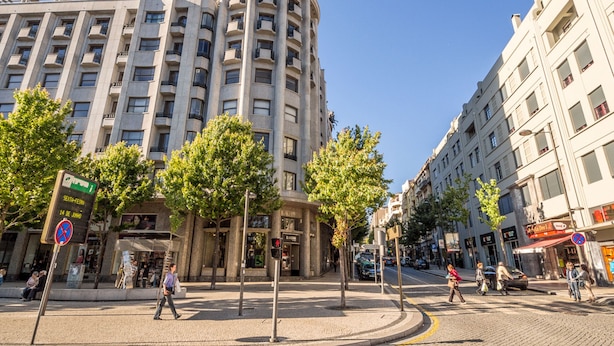 Ein mehrstöckiges Gebäude mit einer gebogenen Ecke, einer Fußgängerampel und Bäumen, die die Straße säumen.