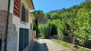 Un edificio con un vialetto sterrato che conduce a un cancello, circondato da vegetazione e un pendio collinare.
