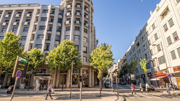 Un immeuble à plusieurs étages doté d’une façade incurvée, une zone piétonne bordée d’arbres et un ciel d’un bleu éclatant.