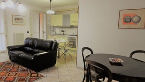 Un salon avec un canapé en cuir noir, une table à manger avec des chaises, un tapis à motifs et, au fond, une cuisine.
