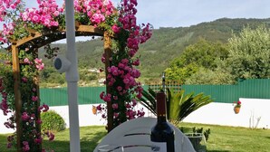 A table with a bottle of wine and two glasses, set under a pergola with pink flowers, overlooking a green landscape.