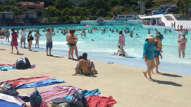 A beach with people swimming and sunbathing, a water slide, and a hill with wind turbines in the background.