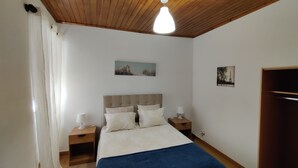 A bedroom with a wooden ceiling, a bed with a blue bedspread, two bedside tables with lamps, and two wall-mounted pictures.