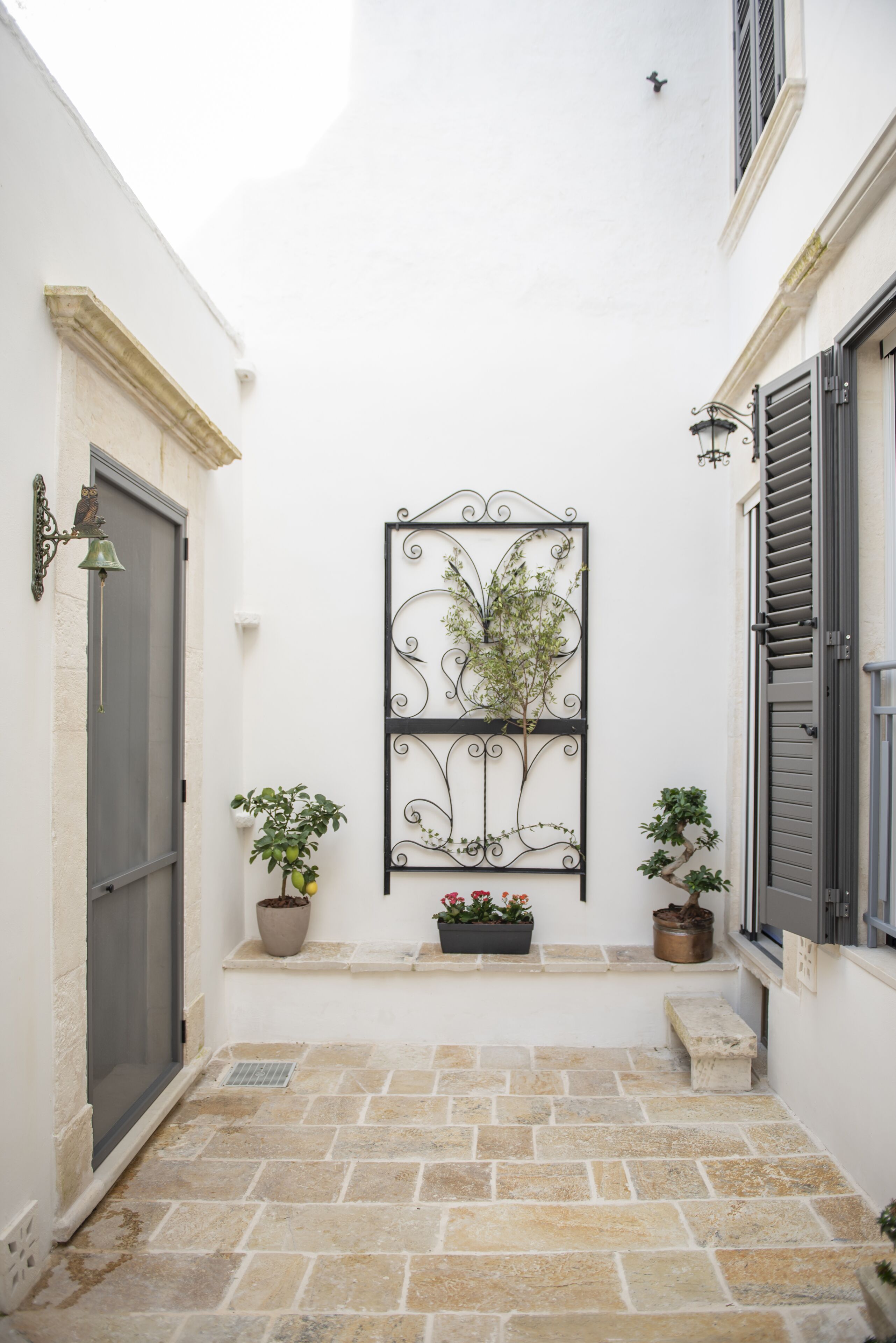 Une cour étroite avec un mur blanc, un portail en métal et des plantes en pot.