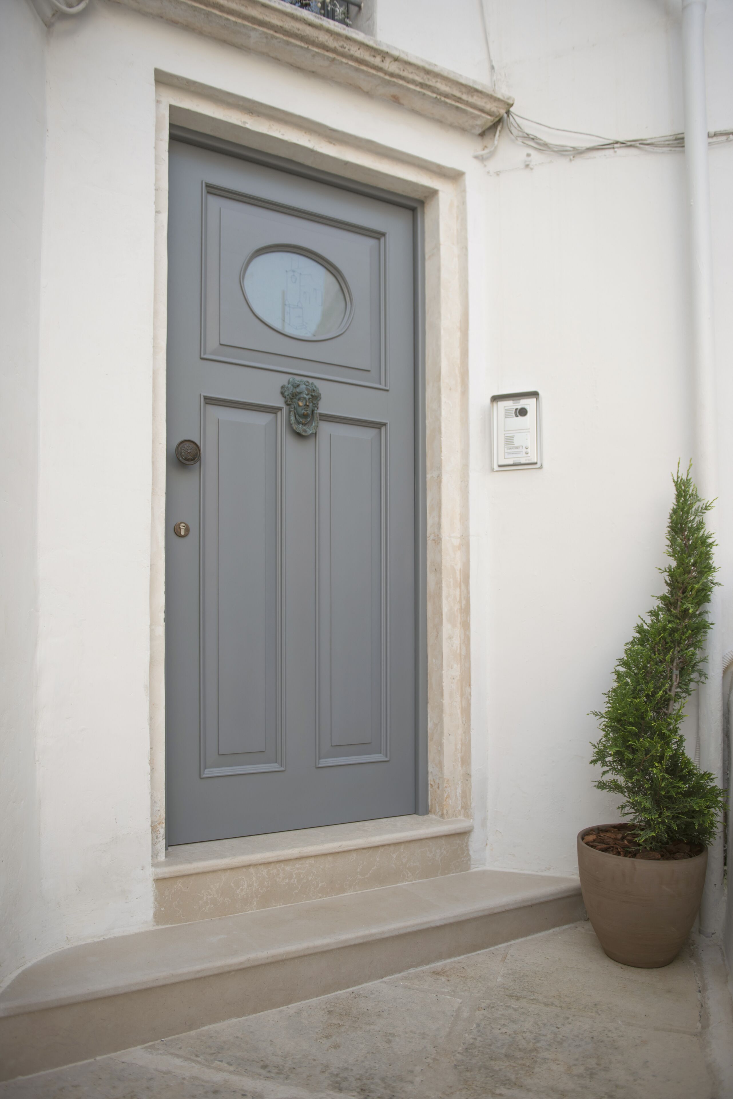 A grey door with a circular window and a decorative knocker, a potted plant beside it, and a small electrical box on the wall.