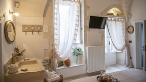 Ein Badezimmer mit einem Steinkopfwaschbecken, einem Spiegel, einem Fenster mit Vorhängen, einem Heizkörper und einem an der Wand befestigten Fernseher.